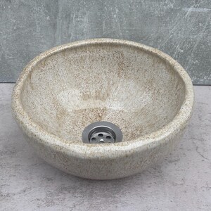 Peut inclure: Un lavabo en céramique beige moucheté de forme ronde. L'intérieur du lavabo est lisse et l'extérieur est texturé. Un drain argenté est visible au fond. Le lavabo est conçu pour une utilisation dans la salle de bain.