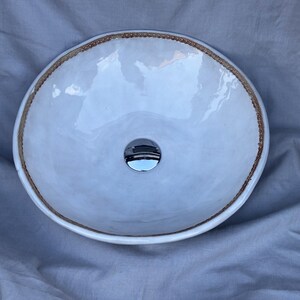 May include: A round, white ceramic sink with a glossy finish. The sink has a decorative, textured brown border and a chrome drain. The sink is designed for bathroom use.