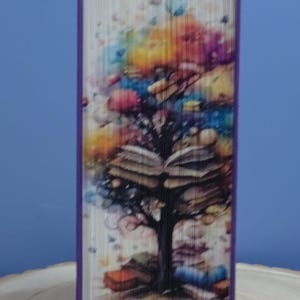 Può includere: Un'opera d'arte in libro piegato con un design ad albero con libri aperti e fogliame colorato. Il libro ha un dorso viola e l'opera è ambientata su uno sfondo blu. Le foglie dell'albero sono un mix di blu, gialli, rosa e viola.