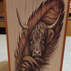 May include: A book art sculpture featuring a folded page design that reveals a detailed image of a Highland cow partially obscured by a large, textured feather. The book has orange covers and white pages, with the artwork printed across the page edges.