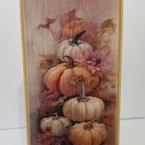 May include: A decorative book with a fall-themed design. The artwork features a stack of pumpkins in shades of orange and white, surrounded by autumn leaves. The book has a gold-colored spine and edges.