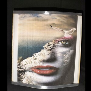 Op de afbeelding: Een ingelijst drukwerk met een surrealistisch beeld van een vrouwen gezicht, met een rotsachtige klif die haar wang en oog vormt. De achtergrond is een bewolkte lucht en een wateroppervlak.