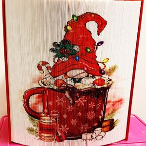 May include: A red and white papercraft gnome wearing a red hat with white snowflakes and lights, holding a red mug with white snowflakes and a brown liquid inside. The gnome is sitting on a green and brown surface with a red and white jar and brown sticks.