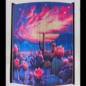 May include: A folded book art piece depicting a desert landscape with a pink and purple sunset, cacti, and blooming flowers.