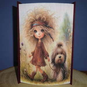 May include: A decorative book with a folded page design featuring a whimsical illustration. The image shows a girl in a brown dress and boots walking a dog on a leash through a field. The book has a burgundy cover.