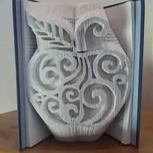May include: A book art sculpture shaped like an apple with intricate cut-out designs. The apple is made from the pages of a book and is a unique and creative piece of art.