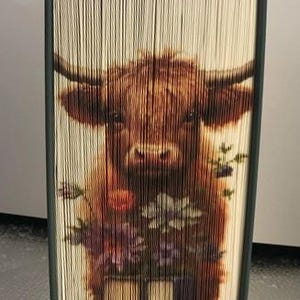 May include: A book art sculpture of a brown Highland cow with a white face, standing in front of a bouquet of purple, pink, and orange flowers. The book is folded into a 3D sculpture.