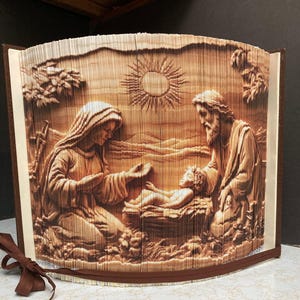 Puede incluir: Una obra de arte en papel marrón que representa una escena religiosa con María, José y el niño Jesús. La obra de arte está hecha de papel plegado, creando un efecto 3D. El sol y el paisaje también son visibles en el fondo. La pieza está encuadernada como un libro con una cinta marrón.