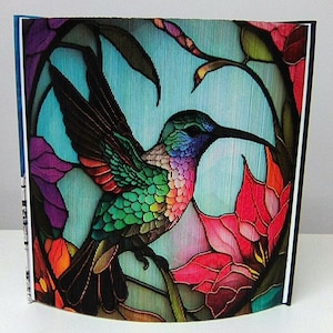 May include: A handcrafted book sculpture featuring a colorful hummingbird design. The bird is depicted with vibrant green, blue, and red hues, surrounded by floral elements. The artwork is set against a light blue background, creating a striking visual effect.