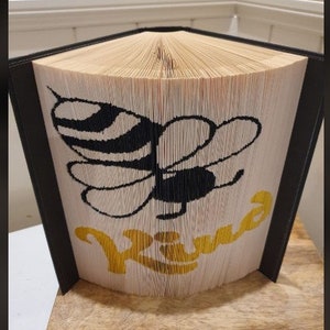 May include: A folded book art piece featuring a black and yellow bee design with the word "Kind" in yellow. The book's pages are folded to create the image, with a black frame and a wooden base.