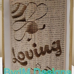 May include: A book art sculpture with the word "loving" cut out of the pages. The sculpture is framed in a wooden frame and has a white background. The artist's signature, "BvdM Designs", is visible at the bottom of the sculpture.