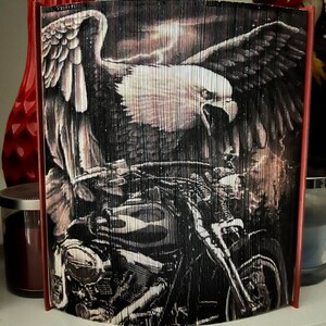 May include: A black and white book art sculpture of an eagle with its wings spread wide, perched above a motorcycle. The eagle is in flight and the motorcycle is in the foreground. The background is a dark, stormy sky.