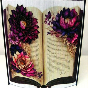 May include: A book art piece with a purple, pink, and white floral design on the cover. The book is open and the pages are folded to create the flowers.