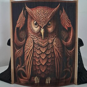May include: A brown and black book sculpture featuring an owl design. The owl is intricately carved into the pages, with detailed feathers and large eyes. The book is propped up by black bookends.