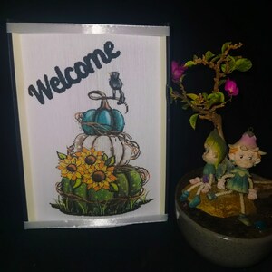 May include: A white card with a black frame and a white ribbon. The card has a black and white illustration of a stack of pumpkins with sunflowers and a bird on top. The card says "Welcome" in black text.