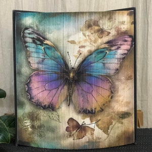 May include: A framed print of a large, iridescent butterfly with blue, purple, and pink wings. The butterfly is set against a background of muted greens and browns, with a smaller butterfly in the lower right corner.