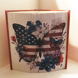 May include: A book art sculpture of a butterfly with the American flag design. The butterfly is surrounded by watercolor flowers in shades of blue, red, and white.