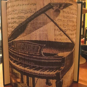 May include: A brown book with a black spine has been folded into the shape of a grand piano. The piano is black with white keys and is open with the lid raised. The book is made of pages from a music book.