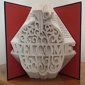 May include: A book folded into the shape of an apple with the word "WELCOME" cut out of the pages. The apple is white and the book is open to reveal the inside of the apple. The book is sitting on a wooden surface with a red background.
