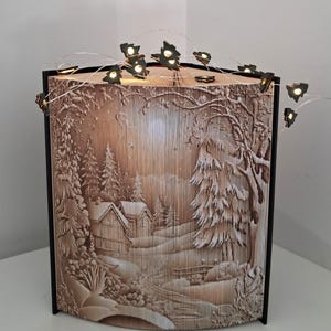 May include: A folded book art sculpture featuring a detailed winter landscape with snow-covered houses and pine trees. The pages are intricately creased to form the scene, and a string of small, illuminated LED lights with tree-shaped clips is draped across the top.