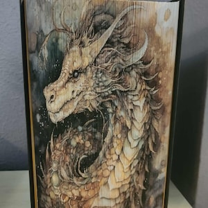 May include: A book art sculpture featuring a detailed dragon illustration printed across the folded pages. The dragon is rendered in shades of brown, tan, and cream with dark accents, showing a side profile of the creature with textured scales.