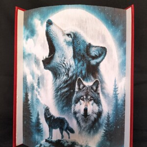 May include: A folded book art piece depicting three wolves howling at the moon. The wolves are in shades of blue and gray, and the background is a dark blue and gray forest scene with a full moon.