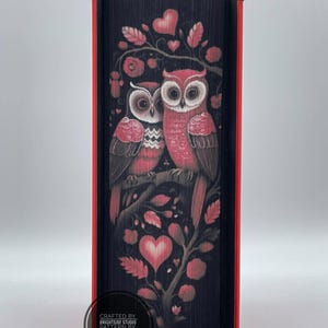 May include: A decorative book with a black cover featuring a design of two owls perched on a branch with hearts and leaves. The owls are red and white, and the book has red edges. The text "CRAFTED BY BRIGHTSIDE STUDIO PATTERN BY BVDM DESIGNS" is printed on the bottom.