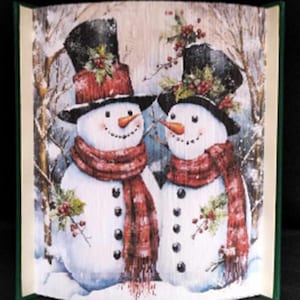 May include: Two snowmen standing side-by-side in a snowy winter scene. The snowmen are wearing black top hats with red and green accents, red scarves, and black buttons. The background is a snowy forest with bare trees.
