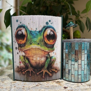 May include: A book art sculpture of a green frog with large, orange eyes. The frog is sitting on a white background with watercolor splatters. The sculpture is made from folded pages of a book.