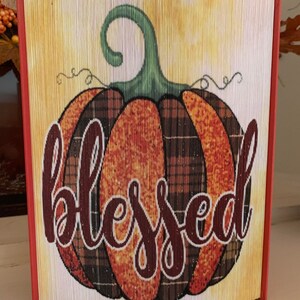 May include: A fall-themed book folding art piece with a large orange pumpkin with a plaid pattern and the word "blessed" in brown cursive lettering.