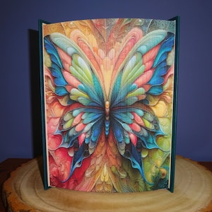 May include: A book sculpture with a vibrant butterfly design. The wings are a mix of blue, green, pink, and orange. The pages are folded to form the butterfly, and the book has a dark green spine. The artwork is displayed on a wooden surface.