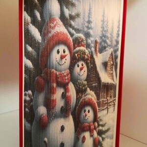 May include: A book art sculpture of three snowmen standing in front of a snowy cabin. The snowmen are wearing red and white hats and scarves. The sculpture is made of folded pages of a book.