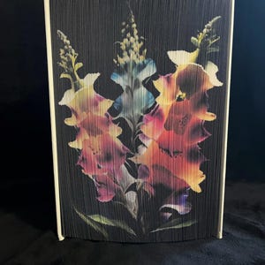 May include: A book art sculpture of three colorful flowers with a black background. The flowers are made of folded pages of a book.