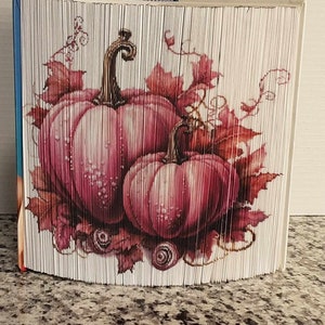 Può includere: Una scultura di arte di libro di tre zucche rosa con steli marroni e foglie in tonalità di marrone, arancio e giallo.
