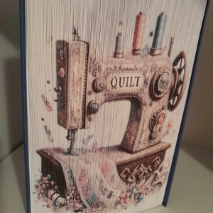 May include: A book art sculpture of a vintage sewing machine with the word "QUILT" on the front. The machine is surrounded by colorful fabric scraps and spools of thread.