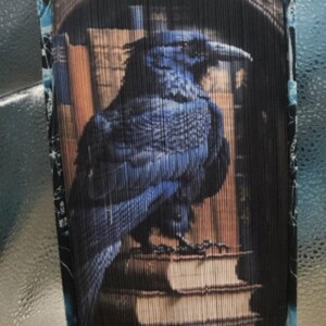 May include: A blue raven perched on a stack of old books, with a dark wooden frame around the image. The raven is facing the right side of the image, with its head turned to the left. The books are stacked in a pile, with the top book being the largest. The frame is made of dark wood and has a simple design.