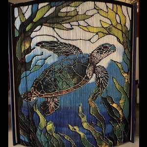 May include: A colorful book art sculpture of a sea turtle swimming in blue water with green seaweed. The sculpture is made of folded pages of a book.