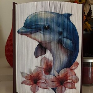 May include: A blue and white folded book art sculpture of a dolphin jumping out of the water with pink flowers.