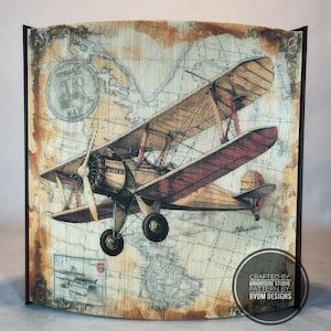 May include: A vintage-style biplane art piece with a map background. The artwork features a detailed illustration of a biplane in shades of brown, red, and beige. The piece is framed in black and includes the text "CRAFTED BY BRIGHTSIDE STUDIO PATTERN BY BVDM DESIGNS".