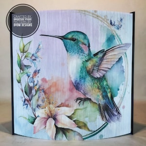 May include: A folded paper art piece depicting a hummingbird in flight with a watercolour style. The hummingbird is green, blue, and purple with a long beak. The background is a soft purple with pink and orange watercolour accents. The hummingbird is perched on a branch with white flowers.