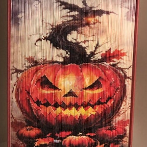 May include: A book art sculpture of a jack-o'-lantern with a menacing grin. The book is folded into a three-dimensional sculpture with a dark background and a spooky, autumnal feel.