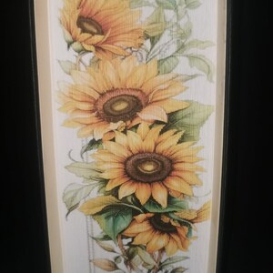 Pode incluir: Um livro com uma capa branca e uma lombada preta. A capa apresenta uma pintura de um buquê de girassóis em um vaso.