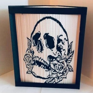 以下が含まれることがあります： 黒い額縁に入った本の芸術作品で、骸骨とバラのデザインが施されています。骸骨は折り畳まれたページで作られており、白黒です。