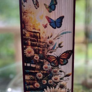 Può includere: Un libro con una copertina di colore bordeaux e una copertina con un design di farfalle colorate. La copertina è realizzata con pagine piegate, creando un effetto 3D. Le farfalle sono disposte in un motivo attorno a un muro di mattoni e fiori.