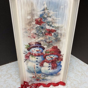 May include: A folded book art sculpture of two snowmen standing in front of a snowy Christmas tree with red poinsettias. The book is decorated with a red ribbon.
