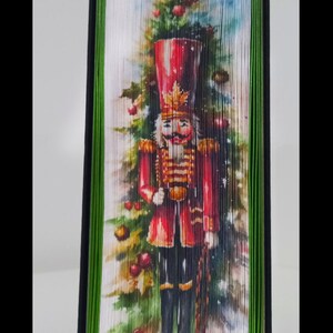 May include: A folded book art piece depicting a nutcracker standing in front of a Christmas tree. The nutcracker is wearing a red and gold uniform and holding a staff. The tree is decorated with baubles and lights. The book is bound in green and black fabric.