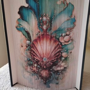 May include: A book art sculpture with a folded book cover featuring a seashell with a pearl inside. The seashell is surrounded by blue, green, and white abstract shapes.