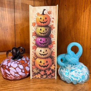 May include: A rectangular book-shaped decor piece featuring a vertical stack of five jack-o'-lanterns surrounded by autumn leaves, flanked by a round purple and orange glass pumpkin and a round light blue and white glass pumpkin with a twisted stem.