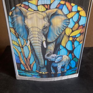 May include: A book art sculpture of an elephant and her calf. The sculpture is made of folded paper and features a stained glass style design with blue, yellow, and orange colors.