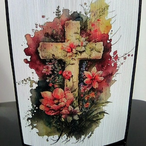 May include: A watercolour painting of a wooden cross surrounded by red and green flowers. The cross is set against a white background with a red and yellow watercolour splatter effect.